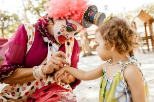 A vibrant clown paints a child's hand at a lively outdoor party, creating a joyful atmosphere.